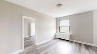 This efficiently designed 2-bedroom, 1-bathroom unit offers a clean, modern aesthetic throughout its single floor. Consistent light-toned hardwood flooring runs through each room, complemented by neutral gray-beige walls and crisp white trim, creating a cohesive and contemporary feel.

The spacious primary bedroom is the standout room of the unit, offering ample square footage with a large window fitted with horizontal blinds that frames lush green outdoor views. A baseboard radiator provides warmth, and a flush-mount ceiling light keeps the space bright. An open doorway connects it to the adjacent room, supporting a flexible layout.

The second bedroom is similarly finished, featuring the same hardwood floors and neutral tones. It includes a generous built-in closet with classic louvered bifold doors, a ceiling fan for added comfort, and its own doorway connecting to the rest of the unit.

The bathroom is clean and functional, featuring a full tub and shower combo with white subway tile surround, chrome fixtures, and a window that allows natural light and ventilation.

The kitchen, visible in the dollhouse model, is neatly positioned at one end of the unit. Throughout, the design prioritizes simplicity and functionality, making efficient use of every area while maintaining a bright, well-maintained atmosphere.