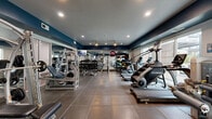 Located at 2901 Indiana St in Dallas, Texas, this fitness center is a comprehensively equipped exercise facility designed to support a full range of workout routines. The space features a bold, modern aesthetic with deep navy blue accent walls contrasting against light grey ceilings, recessed lighting, and pendant-style industrial fixtures that provide ample illumination throughout.

The flooring consists of large-format tile with rubberized matting in high-use areas, offering both durability and safety. Full-length mirrors line the walls, enhancing the sense of openness and allowing users to monitor their form during workouts.

The facility is outfitted with an impressive array of equipment. Cardio offerings include TRUE treadmills, a StairMaster, and elliptical machines positioned along the windowed wall, which allows natural light to filter in and provides views of the surrounding Dallas cityscape. The strength training section houses a Hoist cable/Smith machine system, adjustable benches, and a full dumbbell rack ranging across multiple weight increments. A medicine ball and stability ball station rounds out the functional fitness options.

A wall-mounted flat-screen television provides entertainment, and the open layout ensures comfortable circulation between equipment stations. The overall ambiance is clean, professional, and motivating — well-suited for a serious fitness routine.