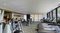 This fitness center offers a comprehensive workout environment designed to accommodate a variety of exercise routines. The space is laid out on a single level, with an open floor plan that allows for comfortable movement between equipment zones. Large floor-to-ceiling windows line one wall, flooding the interior with natural light and creating an airy, expansive atmosphere.

The cardio section is well-stocked with a row of treadmills, elliptical trainers, and additional aerobic machines positioned to take advantage of the natural light from the windows. Strength training equipment, including cable machines and resistance stations, occupies the central area of the floor, providing users with a full range of training options.

The ceiling features a drop-tile design with recessed lighting, while a distinctive purple LED accent strip runs along the upper perimeter of one wall, adding a modern, dynamic ambiance to the space. A mirrored wall further enhances the sense of openness. The floor is finished with patterned commercial carpet tiles that define the space and provide cushioning underfoot, contributing to a functional and professional fitness environment.