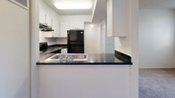 This single-floor unit offers a practical and straightforward layout designed for comfortable everyday living. The living area is carpeted in neutral tones and receives natural light through a window fitted with horizontal blinds, creating a bright and clean atmosphere. A textured popcorn ceiling is consistent throughout the space, adding a classic residential character.

The kitchen is semi-open, separated from the living area by a partial wall with a peninsula counter finished in dark speckled laminate. White cabinetry lines the walls above and below the counter, providing ample storage. Black appliances, including a refrigerator, electric range with overhead ventilation hood, and a stainless steel sink, give the kitchen a defined, functional aesthetic. Light wood-look flooring runs through the kitchen, contrasting with the carpeted areas elsewhere in the unit.

Adjacent to the kitchen, a hallway connects to additional rooms, including what appears to be a bedroom featuring soft carpet and clean white walls. A closet with sliding doors is also present, offering practical storage. The overall design is minimal and neutral, with white walls throughout, creating a blank canvas well-suited for personalization.