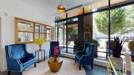Located on NW 18th Avenue in Portland's vibrant Northwest District, this single-floor lobby space offers a thoughtfully designed, contemporary welcome environment. The entry vestibule features polished concrete flooring, a bold sputnik-style chandelier, and a bank of smart parcel lockers, providing both style and practical functionality upon arrival.

Moving deeper into the space, a defining feature is the large-scale, three-dimensional wood mosaic wall installation set against a vivid teal background. Light maple geometric wood pieces are arranged in an abstract, sculptural pattern, creating a striking visual focal point that anchors the interior design aesthetic.

The lounge area is divided into two distinct seating zones. One features richly upholstered navy blue wingback chairs with white piping, arranged around a gold drum coffee table atop a circular woven rug. The second zone offers a more neutral palette with cream-toned armchairs and gold-accented side tables. Both areas benefit from abundant natural light streaming through expansive floor-to-ceiling black-framed glass windows, offering direct views of the tree-lined streetscape outside. Warm wood ceiling accents, pendant lighting with gold interiors, and lush indoor plants complete the refined, welcoming ambiance throughout.