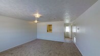 This two-floor property offers a straightforward and functional layout designed for practical everyday living. The main living area is generously sized, featuring wall-to-wall carpet that adds a sense of warmth underfoot, and is framed by clean white walls and a textured ceiling with flush-mounted lighting and HVAC vents. A distinctive architectural detail is the pass-through window cut into the interior wall, connecting the living area to an adjacent room — likely a kitchen or utility space — and providing built-in shelving for added convenience. Toward the front of the home, a tiled entryway leads to a front door fitted with a window panel that draws in natural light, creating a welcoming transition into the main living space. Side windows near the entry further brighten the foyer. The overall aesthetic is simple and neutral, with a light color palette throughout that lends itself well to a variety of furnishing styles. The two-floor layout suggests additional living or sleeping quarters above, providing a clear separation between communal and private areas of the home. This space presents a clean canvas well-suited for personalization and move-in readiness.
