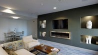 Located at 6010 Attleboro Dr in Cary, NC, this single-floor space showcases a thoughtfully designed interior with a clean, modern aesthetic. The lounge area immediately draws attention with a bold dark accent wall that serves as the focal point of the room, featuring a wall-mounted flat-screen television, recessed display niches, and a sleek linear built-in fireplace with a stainless steel surround. The recessed shelving adds both visual interest and functional display space, contributing to the polished, intentional design of the room. The flooring features a patterned area rug layered over what appears to be a light-toned base floor, adding texture and warmth to the space. Seating is arranged around a rustic wood-and-metal coffee table, creating an inviting, comfortable gathering area. Recessed ceiling lighting provides even, ambient illumination throughout, complementing the room's contemporary palette of neutral whites, grays, and deep greens. The open layout allows for a natural flow between areas, and the overall ambiance is modern and welcoming — well-suited for both relaxed lounging and social interaction.