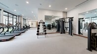 Located at 2358 Bransford Ave in Nashville, TN, this modern fitness center offers a comprehensively outfitted workout facility across a single, expansive floor. The space is divided into two distinct functional zones, each designed to support a full range of exercise modalities.

The strength and functional training area features light hardwood-style flooring and is lined with floor-to-ceiling mirrors that amplify the sense of space and allow users to monitor their form. This zone is equipped with a multi-functional cable and resistance training rig, a full dumbbell rack spanning a wide weight range, medicine balls, and TRX-style suspension trainers — catering to both free weight and functional fitness workouts.

The second zone transitions to plush, light-toned carpet and houses an impressive array of selectorized strength machines and benches, ideal for targeted muscle group training. Large floor-to-ceiling windows flood this area with abundant natural light and offer views of the surrounding outdoor courtyard, creating an open and energizing atmosphere.

A dedicated cardio section features treadmills, ellipticals, a stationary bike with an interactive display, and a stair climber — all positioned to take advantage of the generous window views. Recessed lighting throughout complements the natural light, while exposed concrete columns add an industrial-modern aesthetic to the clean white walls and contemporary finishes.