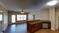 This single-story space showcases an open-concept layout that seamlessly connects the kitchen and living areas, creating a cohesive and functional environment. The kitchen is anchored by a central island topped with dark granite countertops and equipped with a double-basin sink, providing ample workspace and storage through its rich, warm-toned wood cabinetry below. Matching countertops extend along the perimeter of the kitchen, which also includes a dishwasher, contributing to the space's practical design. Hardwood flooring runs throughout, adding warmth and continuity between the kitchen and the adjoining living area. The living space is brightened by large windows, including a set of French doors, which invite abundant natural light and offer views to the outdoors. Crown molding adds a refined architectural detail along the upper walls, while a ceiling fan with a built-in light fixture provides both comfort and illumination. The textured ceiling and neutral-toned walls offer a clean, understated backdrop throughout. A doorway visible from the main space suggests additional rooms beyond, indicating a well-connected and thoughtfully arranged floor plan.