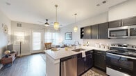 Located at 1500 Laurel Crossing Pkwy NE in Buford, Georgia, this single-floor residence offers a thoughtfully designed open-concept layout with modern finishes throughout. The kitchen serves as a focal point of the home, showcasing dark cabinetry, light granite countertops, a subway tile backsplash, and a center island with a stainless steel sink and pendant lighting. Stainless steel appliances — including a range, microwave, and dishwasher — complete the space. The kitchen flows seamlessly into the living area, which features wood-style flooring, a ceiling fan, and abundant natural light from large windows and a glass-paneled exterior door. The bedroom is a serene retreat with soft neutral tones and direct access to a spacious walk-in closet equipped with wire shelving and carpeted flooring. The bathroom is well-appointed with dark vanity cabinetry, a white countertop, wood-style flooring, and a glass-enclosed tiled shower. A dedicated in-unit laundry closet houses a full-size washer and dryer. Throughout the home, a consistent palette of soft grays, dark fixtures, and warm wood tones creates a cohesive and contemporary aesthetic.