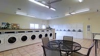 This single-floor laundry facility is thoughtfully designed to accommodate multiple users simultaneously, offering a well-organized and functional layout. The room features a generous row of front-loading washing machines along one wall, complemented by a bank of stacked dryers along the adjacent wall, providing ample capacity for high-volume use. The light yellow walls and large windows allow natural light to brighten the space, creating a welcoming atmosphere. Recessed fluorescent lighting fixtures ensure consistent illumination throughout. Two ceiling fans are mounted overhead, promoting air circulation and comfort during use. The flooring consists of neutral-toned ceramic tiles, which are practical and easy to maintain. A central seating area with a round metal table and mesh chairs offers a comfortable spot for residents to wait. A countertop surface near the washers provides a convenient folding and sorting area. The overall design prioritizes practicality while maintaining a clean, pleasant environment suitable for communal use.
