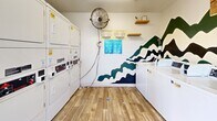 Located at 4341 Spring St in La Mesa, California, this shared laundry facility offers a practical yet visually engaging space designed for everyday convenience. The room is equipped with multiple coin-operated top-load washing machines and stacked washer-dryer units, providing ample capacity for residents. A freestanding utility sink adds further functionality for hand-washing or pre-treating garments.

The interior is finished with durable wood-look plank flooring that lends warmth to the space, while the white walls are elevated by a striking, hand-painted wave mural rendered in deep teal, navy, and white tones — serving as a bold artistic focal point throughout the room. A ceiling-mounted fluorescent light fixture ensures bright, even illumination, and a wall-mounted fan promotes comfortable air circulation.

A large window on one wall invites natural light and provides views of the surrounding outdoor area, creating an open, airy feel. A glass-panel entry door on the opposite side offers additional ventilation and convenient access to the exterior. A waste receptacle is thoughtfully placed within the room for easy use. Posted informational signage and floating wooden shelves round out the space, keeping it organized and user-friendly. Overall, the laundry room balances utility with a clean, cheerful aesthetic.