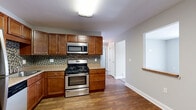 Located at 4543 Camp Rd in Hamburg, NY, this single-floor, two-bedroom, one-bathroom unit offers a practical and well-appointed layout. The kitchen is a standout feature, showcasing warm wood cabinetry, a mosaic tile backsplash, granite-style countertops, and a full suite of stainless steel appliances including a gas range, over-the-range microwave, refrigerator, and dishwasher. Durable hardwood-style flooring runs throughout the kitchen, which connects to the living area via a convenient pass-through window. The living room is comfortably sized with soft carpeting, neutral gray walls, and a large sliding glass door fitted with vertical blinds that allows for abundant natural light. Both bedrooms feature the same plush carpeting and neutral tones, with the primary bedroom offering a spacious closet and a window with natural light. The full bathroom is cleanly finished with large-format tile flooring, a wood vanity with a stone-top sink, and a glass-enclosed shower stall. Throughout the unit, white trim and neutral wall tones create a cohesive, move-in-ready aesthetic.