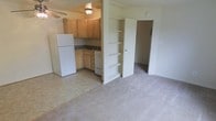 This single-floor unit is designed with an efficient, open-concept layout that seamlessly connects the kitchen and living area. The kitchen is equipped with light wood cabinetry, a white refrigerator, a gas range, and a sink set beneath upper cabinets that provide ample storage. The flooring in the kitchen transitions from a neutral tile to wall-to-wall carpeting that extends throughout the living area, creating a clear visual distinction between the two spaces. A ceiling fan is installed overhead, contributing to the comfort of the main living area. Adjacent to the kitchen, a set of built-in open shelves offers additional storage and display space, adding a practical architectural detail to the unit. A doorway leads to a separate bedroom, which also features carpeted flooring and provides a private retreat from the main living space. The overall color palette is light and neutral, with cream-toned walls and cabinetry contributing to a bright, airy feel throughout. The layout prioritizes functionality and ease of movement, making efficient use of the available space while maintaining a clean, straightforward design throughout.