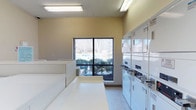 This single-floor laundry facility is designed for practicality and ease of use. The room is illuminated by overhead fluorescent lighting and benefits from natural light through a large, multi-pane window that offers views of the surrounding outdoor greenery. The walls are finished in a warm neutral tone, contributing to a clean and functional atmosphere. Along one side of the room, a row of stacked white washer and dryer units provides ample laundry capacity. A generously sized white folding table runs along the opposite side, offering convenient space for sorting and folding laundry. The flooring is light-toned and easy to maintain, consistent with the room's overall utilitarian design. The textured ceiling adds a modest architectural detail. The space is thoughtfully organized, with enough room to move comfortably between the machines and the folding area. Overall, the facility is well-maintained and efficiently laid out, making it a highly functional shared amenity.