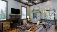 This refined single-floor space showcases a masterfully designed living area that blends traditional elegance with timeless sophistication. The room immediately draws the eye upward to its striking coffered ceiling, finished in rich dark wood tones that contrast beautifully against soft blue-grey walls, creating a sense of architectural depth and grandeur. Recessed lighting is seamlessly integrated into the ceiling's framework, providing warm, layered illumination throughout the space.

A classic fireplace serves as a natural focal point, flanked by a wall-mounted flat-screen television and decorative mantel accents. Opposite the fireplace, a stately vintage-style cabinet with open shelving and ornate detailing offers both display and storage, topped with curated décor including ceramic vessels and a framed landscape painting.

The living area is furnished with plush, neutral-toned sofas and accent chairs arranged to encourage conversation, anchored by a dark coffee table atop stone-look tile flooring. Natural light filters in through a slatted window blind, complementing the ambient glow of a classic table lamp. Sculptural accents, including a dramatic floral arrangement and decorative figurines, add personality and warmth. The overall ambiance is one of curated refinement and comfortable luxury.