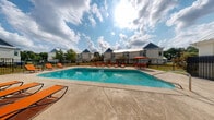 Located at 10532 Klein Rd in Gulfport, Mississippi, this outdoor amenity area centers around a generously sized rectangular swimming pool with clear, blue-tinted water. The pool is surrounded by a broad concrete sundeck that provides ample space for relaxation and recreation. Numerous orange lounge chairs are positioned throughout the deck, offering residents plenty of seating options for sunbathing and leisure. A shaded seating area with an umbrella-covered table and chairs is also available, providing a comfortable spot to unwind out of direct sunlight. The pool area is fully enclosed by black iron fencing, ensuring both security and a clean, cohesive aesthetic. Adjacent to the pool is a single-story pool house featuring white painted brick construction and a metal gabled roof, which houses facilities accessible directly from the deck via an iron-gated entrance. A children's playground is visible in the background, adding to the family-friendly character of the community. The surrounding landscape includes mature trees and well-maintained greenery beneath the open Mississippi sky, creating a bright and inviting outdoor retreat.