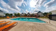 Located at 10532 Klein Rd in Gulfport, Mississippi, this outdoor amenity area centers around a generously sized rectangular swimming pool with clear, blue-tinted water. The pool is surrounded by a broad concrete sundeck that provides ample space for relaxation and recreation. Numerous orange lounge chairs are positioned throughout the deck, offering residents plenty of seating options for sunbathing and leisure. A shaded seating area with an umbrella-covered table and chairs is also available, providing a comfortable spot to unwind out of direct sunlight. The pool area is fully enclosed by black iron fencing, ensuring both security and a clean, cohesive aesthetic. Adjacent to the pool is a single-story pool house featuring white painted brick construction and a metal gabled roof, which houses facilities accessible directly from the deck via an iron-gated entrance. A children's playground is visible in the background, adding to the family-friendly character of the community. The surrounding landscape includes mature trees and well-maintained greenery beneath the open Mississippi sky, creating a bright and inviting outdoor retreat.