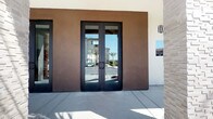 This single-story space makes a strong first impression with its striking exterior entry, framed by sleek black-framed glass double doors set against a rich, dark-stucco façade. The modern entryway offers a glimpse into the bright, open interior beyond.

Inside, the space is defined by a dramatic vaulted ceiling clad in warm wood planking, supported by bold black steel beams that add an industrial contrast to the organic warmth of the wood. Recessed lighting and a cascading pendant chandelier provide layered illumination throughout the open-plan layout.

The main gathering area features a generous communal bar-height table with black upholstered stools, ideal for casual meetings or socializing. A natural stone counter element anchors one side of the space, adding texture and visual interest. Comfortable lounge seating, including cognac leather accent chairs, is arranged throughout.

Toward the rear, large floor-to-ceiling windows and glass doors flood the space with natural light and provide views of the surrounding outdoor areas. The polished tile flooring, crisp white walls, and carefully curated décor — including wall art and fresh floral arrangements — complete the refined, welcoming atmosphere.
