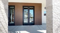 This single-story space makes a strong first impression with its striking exterior entry, framed by sleek black-framed glass double doors set against a rich, dark-stucco façade. The modern entryway offers a glimpse into the bright, open interior beyond.

Inside, the space is defined by a dramatic vaulted ceiling clad in warm wood planking, supported by bold black steel beams that add an industrial contrast to the organic warmth of the wood. Recessed lighting and a cascading pendant chandelier provide layered illumination throughout the open-plan layout.

The main gathering area features a generous communal bar-height table with black upholstered stools, ideal for casual meetings or socializing. A natural stone counter element anchors one side of the space, adding texture and visual interest. Comfortable lounge seating, including cognac leather accent chairs, is arranged throughout.

Toward the rear, large floor-to-ceiling windows and glass doors flood the space with natural light and provide views of the surrounding outdoor areas. The polished tile flooring, crisp white walls, and carefully curated décor — including wall art and fresh floral arrangements — complete the refined, welcoming atmosphere.