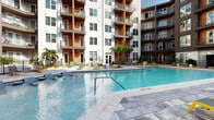 Located at 1601 Central Ave in the heart of Saint Petersburg, Florida, this outdoor amenity space is designed to deliver a resort-like experience within a contemporary urban setting. The centerpiece is a expansive, crystal-clear swimming pool with a shallow sun shelf and in-water lounge areas, surrounded by a spacious stone-tiled deck outfitted with modern yellow-accented chaise lounges and seating. The pool deck is enclosed on multiple sides by the soaring facades of a multi-story residential building, whose warm orange and neutral-toned exteriors, private balconies, and large windows create a dynamic architectural backdrop. Tropical palm trees and lush plantings, including oversized elephant ear foliage, add a distinctly Floridian character throughout the courtyard. A dedicated lounge area features plush white outdoor sofas set on artificial turf, offering a comfortable gathering space adjacent to a vibrant, large-scale botanical mural that injects bold color and artistic energy into the environment. Covered walkways and gated entries connect the various zones of the courtyard, providing both shade and seamless flow. The overall ambiance is modern, inviting, and richly landscaped, reflecting a thoughtfully designed outdoor community space built for the Florida lifestyle.