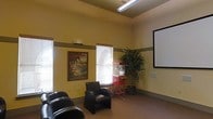 This single-floor communal entertainment space is thoughtfully designed to offer a relaxed and engaging atmosphere for gatherings and movie screenings. The room features warm yellow-toned walls accented by a neutral trim band that adds architectural definition and visual interest throughout the space. Soaring ceilings amplify the sense of openness, while natural light filters in through multiple windows fitted with horizontal blinds, providing a comfortable and adjustable ambiance.

A large wall-mounted projection screen serves as the focal point of the room, making it well-suited for movie nights, presentations, or group viewings. Plush black leather seating is arranged to face the screen, ensuring comfortable sightlines for all occupants. A charming vintage-style red popcorn machine on a classic cart adds a fun, retro character to the space and enhances the entertainment theme.

Decorative touches include a framed vintage travel poster and a lush indoor potted tree, both of which contribute warmth and personality to the environment. Recessed and overhead fluorescent lighting supplements the natural light, ensuring the space remains well-lit at any hour. The combination of functional design, entertainment amenities, and inviting décor makes this a versatile and enjoyable communal venue.