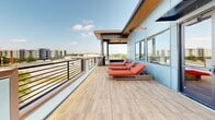 Located at 301 Nichols St in Fort Worth, Texas, this rooftop terrace delivers an exceptional outdoor living experience elevated above the surrounding cityscape. The space is thoughtfully divided into distinct zones, each designed for a specific type of relaxation or entertainment.

A covered lounge area anchors one end of the rooftop, featuring a warm wood-paneled ceiling, ceiling fan, and open-air screened walls that frame panoramic views of the Fort Worth downtown skyline. This section is equipped with a full outdoor kitchen featuring a stainless steel multi-burner gas grill, making it ideal for entertaining. Comfortable wicker sectional sofas with deep red cushions provide ample seating for gatherings.

Transitioning outward, an open sundeck runs the length of the terrace, finished with large-format wood-look porcelain tile. A row of wicker chaise lounges with coral cushions invites sunbathing, while a secondary lounge area with additional seating and a fire pit table offers a more intimate setting.

A covered entertainment nook with tall ceilings, a wall-mounted flat-screen TV, and ceiling fan creates a dedicated outdoor viewing area, perfect for watching games or movies al fresco. Bold blue exterior wall panels and sleek horizontal metal railings give the entire terrace a modern, contemporary aesthetic, while unobstructed views of the Fort Worth skyline and lush tree canopy complete the experience.