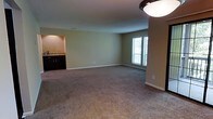 This single-floor residence offers a thoughtfully designed open-concept layout that maximizes the sense of space and ease of living. The main living area is generously proportioned, featuring wall-to-wall plush carpeting that adds warmth and comfort underfoot. Neutral, light-toned walls create a clean and versatile backdrop, complemented by crisp white baseboards that frame the space neatly.

A standout feature is the pass-through kitchen opening, which connects the living area to the kitchen and enhances the open feel of the floor plan. The kitchen is equipped with dark cabinetry and a countertop with a sink visible through the pass-through, offering both functionality and a stylish contrast to the lighter tones of the living space.

Natural light filters into the living area through a large window fitted with horizontal blinds, providing both brightness and privacy control. A flush-mount ceiling light fixture serves as the primary overhead lighting source, ensuring the space remains well-lit.

Overall, the residence presents a practical and comfortable single-level layout with a cohesive design aesthetic — neutral tones, dark cabinetry accents, and soft carpeting — making it a welcoming and functional living environment.