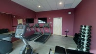 This fitness center is designed to provide a focused and functional workout environment within a single-level layout. The space is immediately defined by its bold deep-red accent walls, which create an energizing atmosphere that complements the purpose of the room. A drop-panel ceiling with recessed lighting ensures the area is well-lit throughout.

The cardio zone is prominently featured, with a row of Matrix-brand treadmills lined up along one wall, each paired with a dedicated wall-mounted flat-screen television — ideal for keeping pace during longer workouts. Additional cardio equipment, including a stationary cycle, is positioned toward the front of the space, offering variety for different training preferences.

The flooring is a light wood-look material that is both durable and easy to maintain, providing a clean and contemporary aesthetic. A small side table near the entrance offers a convenient spot for personal items or refreshments during a session.

The layout is open and uncluttered, allowing for comfortable movement between equipment. A single entry door provides straightforward access to the room. A wall-mounted speaker is also present, supporting an audio-enhanced workout experience. Overall, this fitness center is a practical and well-appointed space suited for daily cardiovascular and general exercise routines.