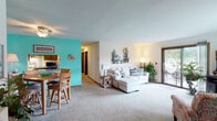 This single-floor residence offers a thoughtfully arranged layout that balances comfort and functionality throughout. The open-concept living and dining area forms the heart of the home, where soft carpeting, neutral walls, and abundant natural light from a sliding glass door create a welcoming atmosphere. The sliding door leads directly to a private balcony with pleasant outdoor views, seamlessly blending indoor and outdoor living. The dining area makes a bold statement with vibrant teal accent walls, a ceiling fan, and space for a full dining table — flowing naturally into the adjacent kitchen. The kitchen is efficiently designed with warm oak cabinetry, tile flooring, ample counter space, and stainless steel appliances, including a full-size refrigerator. A second sitting area near a large window provides an additional relaxing retreat filled with natural light. The two bedrooms are carpeted and comfortable, each featuring ceiling fans and generous closet space with mirrored sliding doors. The bathrooms are finished with oak vanity cabinetry, light countertops, tile flooring, and large mirrors. A consistent coastal-inspired aesthetic runs throughout the space, contributing to a calm and cohesive ambiance across every room.