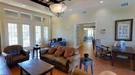 This single-story community space combines comfort and functionality in a warm, inviting setting. The main lounge area draws the eye upward with soaring ceilings and an elegant wrought-iron chandelier fitted with a classic globe shade, casting a soft glow over the room. Large French doors line the walls, flooding the interior with abundant natural light and offering views of the surrounding outdoor areas. Terra cotta tile flooring runs throughout, reinforcing the space's Mediterranean-inspired character.

The lounge is furnished with rich brown leather sofas accented by warm-toned throw pillows, complemented by dark wood side tables and accent chairs with ornate upholstery — creating a refined yet approachable atmosphere. A reception or administrative desk equipped with a computer workstation is positioned near the rear of the lounge, indicating the space's role as a staffed community hub.

Through a set of glass-paned French doors, a well-equipped fitness room is visible, featuring exercise machines and ample natural light from its own windows. The overall ambiance is one of understated elegance, blending practical amenities with tasteful décor to create a welcoming environment suited for both social gatherings and everyday community use.