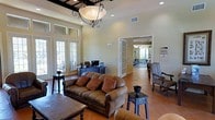 This single-story community space combines comfort and functionality in a warm, inviting setting. The main lounge area draws the eye upward with soaring ceilings and an elegant wrought-iron chandelier fitted with a classic globe shade, casting a soft glow over the room. Large French doors line the walls, flooding the interior with abundant natural light and offering views of the surrounding outdoor areas. Terra cotta tile flooring runs throughout, reinforcing the space's Mediterranean-inspired character.

The lounge is furnished with rich brown leather sofas accented by warm-toned throw pillows, complemented by dark wood side tables and accent chairs with ornate upholstery — creating a refined yet approachable atmosphere. A reception or administrative desk equipped with a computer workstation is positioned near the rear of the lounge, indicating the space's role as a staffed community hub.

Through a set of glass-paned French doors, a well-equipped fitness room is visible, featuring exercise machines and ample natural light from its own windows. The overall ambiance is one of understated elegance, blending practical amenities with tasteful décor to create a welcoming environment suited for both social gatherings and everyday community use.
