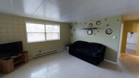 This single-floor space presents a modest and functional living area defined by its distinctive block-tile wall construction, finished in a muted green tone that lends a calm, understated character to the room. The ceiling features a clean, paneled white finish that brightens the interior and contributes to an open feel. Light enters the space through a window fitted with horizontal blinds, offering a view of mature trees outside and providing a natural connection to the outdoors. The flooring consists of light-toned vinyl tiles, which are easy to maintain and reflect ambient light throughout the room. A baseboard heating unit runs along the exterior wall, ensuring efficient climate control. The accent wall showcases playful circular decal artwork alongside a wall-mounted shelf and clock, adding personality and visual interest to the space. The layout is straightforward and practical, allowing for flexible furniture arrangement. Overall, the space offers a functional, low-maintenance environment with a distinctive character rooted in its architectural finishes and simple, utilitarian design.
