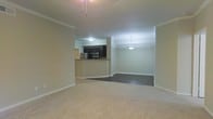 This single-floor space showcases an open-concept design that seamlessly connects the living, dining, and kitchen areas. The living area features plush, neutral-toned carpet and is adorned with crown molding along the ceiling, adding a refined architectural detail to the space. A ceiling fixture with pull chains provides overhead lighting in this zone.

The kitchen is positioned at the back of the space and is defined by dark cabinetry, granite or stone countertops, and stainless steel appliances, including a refrigerator and microwave. A raised breakfast bar serves as a natural divider between the kitchen and the adjoining areas, offering casual seating potential and a functional workspace.

Adjacent to the kitchen, the dining area transitions to rich, dark hardwood flooring, creating a clear visual distinction between zones. A pendant light fixture hangs above this space, providing focused illumination and adding an elegant touch. The neutral, light-toned walls throughout the space contribute to a bright and cohesive ambiance, while the open layout enhances the overall sense of flow and spaciousness.
