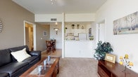 This single-floor residence is designed around an open-concept floor plan that seamlessly connects the living room, dining area, and kitchen into one cohesive living space. The living room is carpeted in a warm neutral tone and is anchored by a wood-framed glass coffee table and a leather sofa, providing a comfortable and functional gathering space. A half-wall with a pass-through counter separates the kitchen from the main living area while maintaining visual openness. The kitchen features white cabinetry, a full-size refrigerator, and a tiled backsplash, with decorative greenery displayed on top of the upper cabinets adding a touch of personality. The dining area sits just beyond the living room, with hardwood-style flooring, a wooden dining table and chairs, and an overhead pendant light fixture that adds warmth to the space. The walls throughout are painted in soft, neutral tones, complemented by white trim and standard ceiling heights that contribute to a clean and airy atmosphere. Recessed and track lighting provide consistent illumination across the open layout. Hallways leading from the main living area suggest additional private rooms beyond, rounding out this well-organized and efficiently designed single-level home.