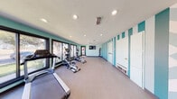 Located at 6501 Eastridge Rd in Odessa, Texas, this fitness center is a thoughtfully designed single-floor space that offers a comprehensive workout environment. The room is decorated in a refreshing palette of soft mint green and bold teal, with eye-catching vertical striped and geometric accent walls that lend the space a modern, energetic ambiance.

The facility is generously equipped with multiple treadmills and elliptical machines positioned along a wall of large, floor-to-ceiling windows that flood the space with natural light and provide pleasant outdoor views of the surrounding greenery. A full-length mirror spans one wall, visually expanding the room while allowing users to monitor their form during exercise.

Additional equipment includes a medicine ball rack stocked with a variety of weighted balls for functional training, as well as strength training machines. A wall-mounted flat-screen TV is positioned for convenient viewing during workouts. Recessed lighting throughout the ceiling ensures the space remains bright and well-lit at all times.

The flooring is durable and fitness-appropriate, providing comfort underfoot across the open workout area. The layout is spacious and uncluttered, allowing for ease of movement between stations. A direct exterior door provides convenient access to and from the outdoors, rounding out this functional and inviting fitness facility.