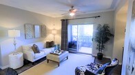 This single-floor space showcases a well-appointed living room designed for both comfort and style. The room features crown molding along the ceiling, adding a refined architectural detail to the space. A ceiling fan with an integrated light fixture serves as a functional centerpiece overhead.

Natural light fills the room through a large sliding glass door fitted with floor-to-ceiling curtain panels, providing direct access to a private balcony with outdoor views of surrounding greenery. The combination of hard flooring and a plush area rug defines the seating area, offering textural contrast underfoot.

The living space is furnished with a white sofa accented by decorative pillows, complemented by a tufted ottoman that doubles as a coffee table. A bold patterned accent chair adds visual interest to the arrangement. Wall art panels and a tall potted indoor plant contribute to the overall aesthetic, bringing a natural element indoors.

Warm ambient lighting from a floor lamp enhances the inviting atmosphere during evening hours. The neutral wall tones provide a versatile backdrop, while curated décor in teal, brown, and white tones create a cohesive and modern interior feel throughout the space.