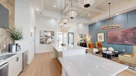 Located on Browning Way in Foley, Alabama, the Exchange at Foley clubhouse is a thoughtfully designed single-floor community space that blends coastal charm with contemporary sophistication. The open-plan interior is anchored by a striking coffered ceiling with exposed wood beam detailing, recessed lighting, and an array of pendant globe fixtures that cast warm, ambient light throughout. Light-toned hardwood floors run seamlessly across the entire space, enhancing its bright and airy character. A prominent white quartz reception and presentation counter serves as the focal point upon entry, complemented by a nearby refreshment bar with a herringbone wood accent wall, mounted flat-screen television, and bar seating. The lounge area features a richly colored blue-gray wainscoted accent wall adorned with curated artwork, anchoring a comfortable seating arrangement beneath a wrought-iron chandelier. Large floor-to-ceiling windows and glass doors on the rear wall flood the space with natural light and provide direct views of the outdoor pool area. Decorative drapery adds texture and warmth, while an eclectic selection of furnishings and artwork contributes to the space's vibrant, welcoming atmosphere—making it an ideal gathering place for residents and visitors alike.