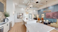 Located on Browning Way in Foley, Alabama, the Exchange at Foley clubhouse is a thoughtfully designed single-floor community space that blends coastal charm with contemporary sophistication. The open-plan interior is anchored by a striking coffered ceiling with exposed wood beam detailing, recessed lighting, and an array of pendant globe fixtures that cast warm, ambient light throughout. Light-toned hardwood floors run seamlessly across the entire space, enhancing its bright and airy character. A prominent white quartz reception and presentation counter serves as the focal point upon entry, complemented by a nearby refreshment bar with a herringbone wood accent wall, mounted flat-screen television, and bar seating. The lounge area features a richly colored blue-gray wainscoted accent wall adorned with curated artwork, anchoring a comfortable seating arrangement beneath a wrought-iron chandelier. Large floor-to-ceiling windows and glass doors on the rear wall flood the space with natural light and provide direct views of the outdoor pool area. Decorative drapery adds texture and warmth, while an eclectic selection of furnishings and artwork contributes to the space's vibrant, welcoming atmosphere—making it an ideal gathering place for residents and visitors alike.