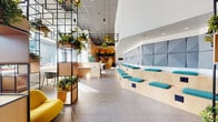 Located at 3333 S La Cienega Blvd in Los Angeles, this thoughtfully designed two-floor space blends contemporary architecture with biophilic elements to create a vibrant, welcoming environment. The ground floor makes an immediate impression with its dramatic double-height lobby, where towering black metal grid structures adorned with cascading greenery and suspended planters draw the eye upward. Large floor-to-ceiling windows flood the space with abundant natural light, while large-format gray tile flooring anchors the expansive open layout. A stepped tiered seating platform lined with teal cushions and geometric acoustic wall panels provides a functional and visually engaging gathering area. Teal lounge chairs, yellow accent seating, and curated abstract artwork contribute to the space's energetic yet refined palette. A fully equipped kitchen features bold teal cabinetry, stainless steel fixtures, a built-in tap, and a large window that connects the interior to the surrounding cityscape. The second floor offers a more intimate lounge setting, distinguished by warm light wood flooring, a striking diagonal-slat wood accent wall, a navy sofa, plush ottomans, and a glass-railed mezzanine overlooking the lobby below. Throughout both floors, lush indoor plants reinforce the space's commitment to a nature-inspired design philosophy.