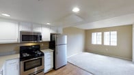 This single-floor residence offers a clean, functional layout centered around an open-concept kitchen and living area. The kitchen is outfitted with stainless steel appliances, including a full-size refrigerator, electric range, and over-the-range microwave, all complemented by crisp white cabinetry and light granite-style countertops. The cabinetry provides ample storage, while the layout ensures efficient use of space for everyday cooking and meal preparation. The kitchen flows seamlessly into the living area, which features a combination of hardwood-style flooring and plush carpeting, creating distinct zones for movement and relaxation. Neutral beige walls keep the space feeling warm and versatile, suitable for a variety of furnishing styles. A window in the living area invites natural light, brightening the room and contributing to an open, airy feel. Recessed lighting and a flush ceiling fixture provide consistent illumination throughout. The overall ambiance is practical and comfortable, with modern finishes and a straightforward design that prioritizes livability. The cohesive color palette and updated appliances make this a move-in-ready space well suited for everyday living.