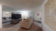 This single-floor residence offers a thoughtfully arranged layout that maximizes comfort and functionality across its key living spaces. The living room serves as the central gathering area, featuring wall-to-wall carpeting in warm neutral tones and a large mirrored wall accent that visually expands the room. Vertical blinds along the exterior-facing wall provide light control, while the open flow connects seamlessly to a dining area nearby. The kitchen is efficiently designed with ample white cabinetry, dark countertops, white appliances including a range, dishwasher, and refrigerator, and easy-to-maintain tile flooring. Storage is well-distributed throughout the cabinetry. The bedroom is generously proportioned, accommodating a full bedroom suite with room to spare. Soft carpeting continues here, and a window dressed with layered blue curtains allows natural light to filter in. The bathroom features classic tile wainscoting, a medicine cabinet with mirror, a single vanity, and a tub-shower combination. Throughout the unit, the consistent use of neutral tones and practical finishes creates a cohesive, livable environment.