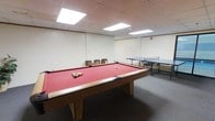 This game room is a dedicated recreational space designed for leisure and social interaction. The room's centerpiece is a full-size pool table with a striking red felt surface and a solid wood frame, fully racked and ready for play. Adjacent to the pool table, a regulation-size ping-pong table provides an additional activity option for guests. A set of cushioned stools is positioned along the wall, offering convenient seating between games. The room features a drop ceiling with recessed panel lighting that provides bright, even illumination throughout the space, ensuring comfortable visibility for all activities. The neutral white walls and carpeted flooring create a clean, functional environment. The layout provides ample clearance around both tables, allowing players to move freely and comfortably during use. The room's straightforward design keeps the focus on its recreational offerings, making it a practical and well-equipped space for entertainment.