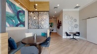 Located at 2810 University Square Drive in Tampa, Florida, this thoughtfully designed single-floor amenity space blends contemporary aesthetics with practical functionality. Light-toned hardwood flooring flows throughout the open common areas, complemented by neutral walls adorned with striking curated artwork, decorative wall sculptures, and mixed-media installations that add visual depth and character.

The lounge and dining area features a mix of cozy booth seating and freestanding tables paired with modern chairs, all illuminated by statement pendant lighting with warm Edison-style bulbs. A wood-accented ceiling detail frames this gathering space, lending a refined warmth to the atmosphere. Floor-to-ceiling windows dressed with teal-patterned drapes invite natural light and provide views of the surrounding greenery.

Sliding barn-style doors open into a versatile conference room furnished with a richly grained dark wood table, wall-mounted display screen, and elegant decorative accents including a sunburst mirror and botanical artwork. A dedicated business center offers built-in workstations with desktop computers set against a bold teal and white geometric accent wall, ideal for productivity.

Throughout, connecting corridors feature decorative ring and moon-shaped wall sculptures, creating a cohesive, gallery-like aesthetic. The overall ambiance is modern, vibrant, and welcoming.