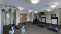 This single-floor fitness center offers a functional and organized workout environment designed to meet a variety of exercise needs. The space is outfitted with a selection of cardio and strength training equipment, including a treadmill, an elliptical machine, a stationary bike, and a multi-use cable weight machine, providing a well-rounded workout experience. A wall-mounted television is positioned near the cardio equipment, allowing users to stay entertained during their sessions. The flooring features a durable, dark carpet with rubber exercise mats placed beneath the larger equipment for added stability and floor protection. The room is illuminated by flush-mount ceiling light fixtures that provide even, practical lighting throughout. The textured ceiling and neutral-toned walls give the space a clean, no-frills aesthetic focused on utility. A set of double wooden doors with automatic closers serves as the main entry point, and the layout allows for comfortable movement between stations. Overall, the fitness center is a straightforward, purpose-built space that delivers essential amenities for maintaining an active routine.