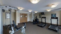 This single-floor fitness center offers a functional and organized workout environment designed to meet a variety of exercise needs. The space is outfitted with a selection of cardio and strength training equipment, including a treadmill, an elliptical machine, a stationary bike, and a multi-use cable weight machine, providing a well-rounded workout experience. A wall-mounted television is positioned near the cardio equipment, allowing users to stay entertained during their sessions. The flooring features a durable, dark carpet with rubber exercise mats placed beneath the larger equipment for added stability and floor protection. The room is illuminated by flush-mount ceiling light fixtures that provide even, practical lighting throughout. The textured ceiling and neutral-toned walls give the space a clean, no-frills aesthetic focused on utility. A set of double wooden doors with automatic closers serves as the main entry point, and the layout allows for comfortable movement between stations. Overall, the fitness center is a straightforward, purpose-built space that delivers essential amenities for maintaining an active routine.