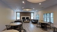 This single-floor space functions as a versatile communal lounge, thoughtfully designed to accommodate both relaxed socializing and casual dining. The room's most notable architectural feature is a circular tray ceiling detail at its center, housing a flush-mount light fixture surrounded by recessed lighting that casts an even, welcoming glow throughout the space. A suspended tile ceiling with additional recessed lights extends across the remainder of the room, maintaining a clean and uniform aesthetic.

The flooring is composed of light-toned wood-look planks, lending warmth and continuity to the open layout. The walls are finished in a neutral palette, with a rich brown accent wall serving as a focal point along the back. This accent wall frames a decorative fireplace mantel flanked by two windows dressed with horizontal blinds, allowing natural light to filter in while maintaining privacy.

The furnishings are arranged to support multiple uses: a seating area features dark leather sofas and armchairs grouped around a low coffee table, while round dining tables with dark metal chairs occupy the perimeter, offering flexible gathering options. Wall sconces add an additional layer of ambient lighting, contributing to the space's inviting, lounge-like atmosphere.