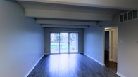 This single-story space presents a clean, open living area defined by rich, dark hardwood flooring that runs the length of the room, reflecting natural light and adding warmth to the interior. The ceiling features a prominent exposed structural beam that spans the width of the space, lending an architectural character to the room. The walls are finished in a cool, neutral blue-gray tone, creating a calm and contemporary atmosphere throughout.

A central sliding glass door serves as the focal point of the main living area, fitted with vertical blinds and framed by views of a well-maintained outdoor green space. This feature allows abundant natural light to flood the interior, enhancing the sense of openness. HVAC vents are positioned along the upper walls, ensuring efficient climate control throughout the space.

White baseboards provide a crisp contrast against both the darker flooring and the wall color, contributing to a polished finish. An arched entryway is visible to one side, hinting at additional rooms beyond the main living area. The overall ambiance is spacious, airy, and move-in ready, with a modest yet stylish aesthetic that balances functionality and comfort.