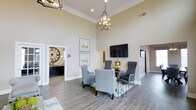 Located on State Street, this single-story space makes a strong first impression with its dramatic soaring ceilings and refined interior design. The main living area serves as the focal point, anchored by a seating arrangement of upholstered gray accent chairs positioned around a central side table — an inviting setup suited for conversation or leisure. Overhead, a geometric open-frame chandelier provides a stylish focal point, complementing the recessed lighting that illuminates the room evenly.

The walls are finished in a warm neutral tone, enhanced by crisp crown molding that adds architectural character throughout. Wide-plank hardwood-style flooring runs across the space, contributing warmth and continuity. A wall-mounted flat-screen television is positioned above a small decorative accent, making the area functional as well as aesthetically polished.

An adjoining room, visible through a wide doorway, features a large decorative wall clock and darker cabinetry, suggesting a dedicated office or concierge area. Additional passageways hint at further rooms beyond, offering a sense of depth and connectivity throughout the layout. Overall, the space exudes an elegant, welcoming ambiance.