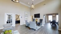Located on State Street, this single-story space makes a strong first impression with its dramatic soaring ceilings and refined interior design. The main living area serves as the focal point, anchored by a seating arrangement of upholstered gray accent chairs positioned around a central side table — an inviting setup suited for conversation or leisure. Overhead, a geometric open-frame chandelier provides a stylish focal point, complementing the recessed lighting that illuminates the room evenly.

The walls are finished in a warm neutral tone, enhanced by crisp crown molding that adds architectural character throughout. Wide-plank hardwood-style flooring runs across the space, contributing warmth and continuity. A wall-mounted flat-screen television is positioned above a small decorative accent, making the area functional as well as aesthetically polished.

An adjoining room, visible through a wide doorway, features a large decorative wall clock and darker cabinetry, suggesting a dedicated office or concierge area. Additional passageways hint at further rooms beyond, offering a sense of depth and connectivity throughout the layout. Overall, the space exudes an elegant, welcoming ambiance.