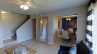 This two-story home offers a thoughtfully designed main level where the living, dining, and kitchen areas flow seamlessly into one another, creating an open and functional layout. The main living area features light tile flooring, neutral gray walls, and a ceiling fan overhead, lending a clean and airy feel to the space. A full-length mirror with a rustic wood frame adds a decorative touch while amplifying the sense of space. A staircase with a dark wood railing leads to the upper level, which houses additional living quarters. The dining area is a standout feature, distinguished by a bold navy blue accent wall adorned with circular wall art. A wood dining table surrounded by upholstered chairs sits beneath a classic chandelier, creating an elegant yet inviting atmosphere. Adjacent to the dining space, the kitchen is partially visible, offering white cabinetry and bright lighting. The cohesive design language throughout the main level — combining neutral base tones with carefully chosen accent elements — gives the home a polished, contemporary character. The open sightlines between rooms enhance the sense of connectivity and make the space feel welcoming and well-suited for both everyday living and entertaining.