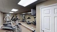 This dedicated fitness space is designed to provide a comprehensive workout environment within a single, efficiently organized room. The space is outfitted with a range of exercise equipment, including a treadmill, elliptical trainer, stationary bike, and a multi-function cable/weight machine, offering users the ability to engage in both cardiovascular and strength-training routines without leaving the premises.

A defining architectural feature of the room is the full-length wall mirror that spans the length of the exercise area, creating a sense of expanded depth while allowing users to monitor their form during workouts. The drop ceiling is fitted with large, recessed fluorescent light panels that ensure the room is evenly and brightly illuminated throughout. A wall-mounted television is positioned for easy viewing during exercise sessions, adding a layer of convenience and entertainment.

The neutral, taupe-toned walls and light-colored flooring contribute to a clean and focused atmosphere. A wall-mounted climate control unit ensures the space remains comfortable during intensive workouts. The room is accessed through a standard interior door, keeping it self-contained and functional. Overall, the space is thoughtfully arranged to maximize utility and comfort for a private fitness environment.