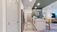 This one-bedroom, one-bathroom residence is designed to maximize efficiency without sacrificing style. The open-concept kitchen and living area serves as the heart of the space, where a galley-style kitchen flows seamlessly into the living room. The kitchen is outfitted with white shaker cabinetry, stone-topped counters, stainless steel appliances, and a peninsula island with bar seating — ideal for casual dining or entertaining. A bold teal accent wall adds a striking visual contrast to the otherwise bright, neutral palette. Light wood-toned flooring extends throughout the main living area, complementing the airy, modern aesthetic.

The living room offers natural light through large windows dressed with floor-length drapes, a ceiling fan overhead, and ample space for comfortable furnishings. A hallway connects the main living area to the bedroom and bathroom, with a dedicated closet providing storage.

The bedroom features plush carpeting, natural light from a window with exterior views, a ceiling fan, and a distinctive arched accent wall detail. The bathroom includes a tiled tub-shower combination and clean, neutral finishes throughout.