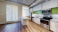 This main floor unit at 10620 116 Street NW in Edmonton showcases a thoughtfully designed industrial-modern aesthetic throughout. The open-concept living and kitchen area is defined by striking exposed concrete ceilings paired with track lighting, which together create an urban loft ambiance. Wood-look flooring runs consistently throughout the unit, adding warmth and visual continuity that complements the raw architectural elements overhead.

The kitchen is a standout feature, outfitted with sleek white cabinetry extending to the ceiling, a bold green glass backsplash, and a full suite of stainless steel appliances including a refrigerator, electric range, built-in dishwasher, and over-the-range microwave. A central island with a light grey countertop provides additional workspace and casual dining potential. Natural light filters into the living area through a large window dressed with roller and vertical blinds.

The bedroom is a calm, neutral space with the same warm flooring and exposed concrete ceiling, served by a ceiling fan and a frosted-glass sliding door for privacy. The bathroom is clean and modern, featuring large-format white wall tiles surrounding a soaker tub and shower combination, wood-look tile flooring, and a minimalist vanity with a grey countertop. A utility area with an electrical panel and storage access is efficiently tucked within the unit.